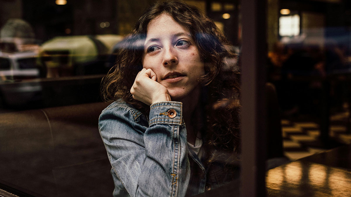 Young woman in a denim jacket looking thoughtfully out a window, reflecting on things people used to spend hours doing