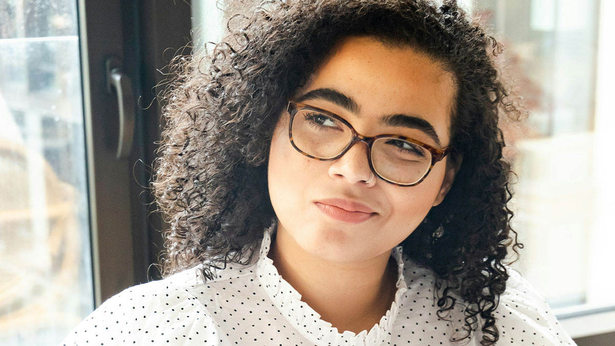 Young woman with curly hair and glasses looking thoughtfully by a window, illustrating the importance of sleep for well-being.