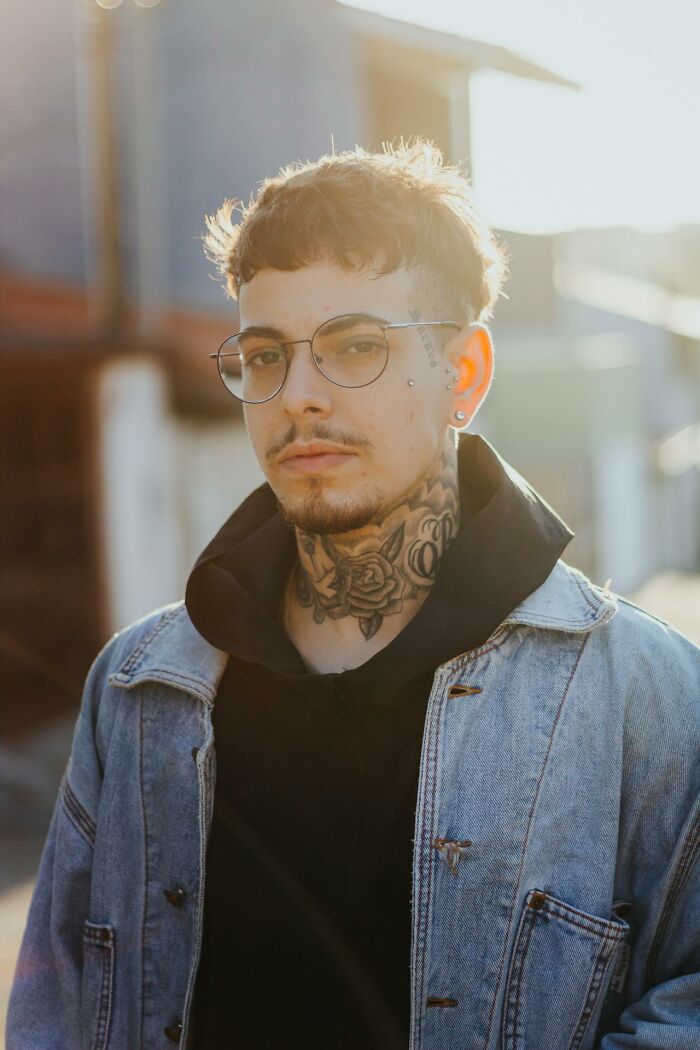 Young man with neck and face tattoos wearing glasses and a denim jacket, showcasing tattoo art requested by clients.
