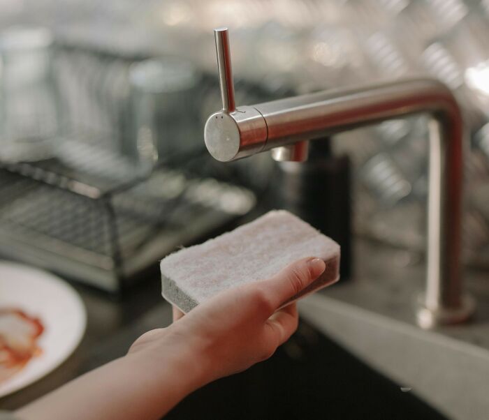 Hand holding a sponge under faucet in kitchen sink, illustrating things people used to spend hours doing pointlessly.