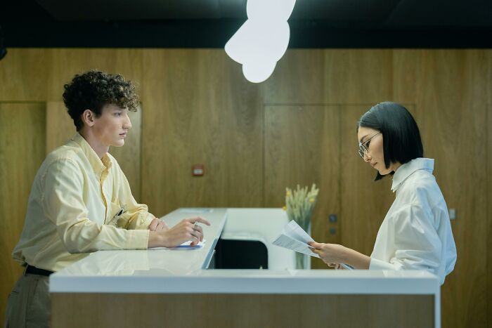 Young man and woman at a reception desk, the scene subtly hinting at hidden manipulation and betrayal.