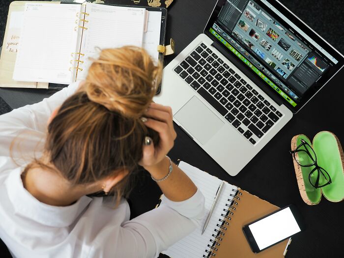 Overhead view of a stressed person at a desk with laptop, planner, and phone, representing wasted hours on pointless tasks.