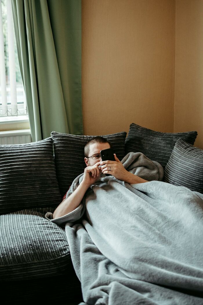 Man relaxing on a couch covered with a blanket, using a phone in a cozy living room setting, highlighting gender freedom topics.