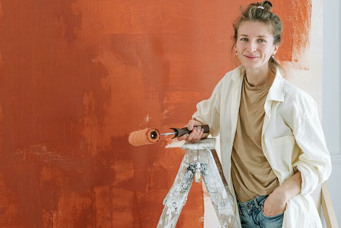 Woman holding paint roller, standing by ladder while painting wall, illustrating subtle ways misogyny affects daily life.