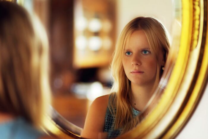Young woman looking thoughtfully at herself in a round mirror reflecting wasted hours spent on pointless activities.