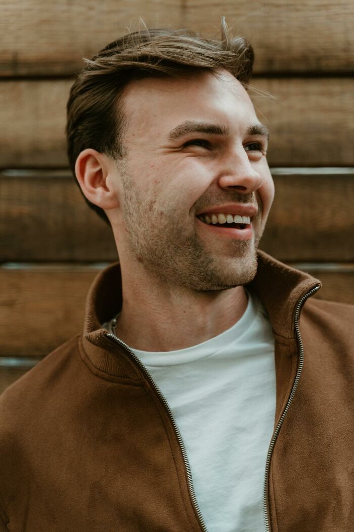 Young man smiling and looking away, wearing a brown jacket and white shirt with a wooden background, tattoo client concept.