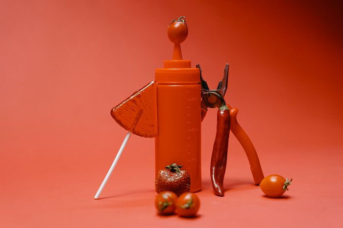 Orange quirky objects including a bottle, lollipop, pliers, chili pepper, and tomatoes arranged against an orange background. Orange quirky objects including a bottle, lollipop, pliers, chili pepper, and tomatoes arranged against an orange background.