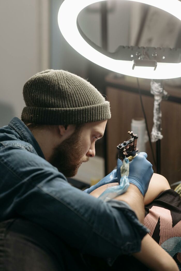 Tattoo artist wearing a beanie and gloves focused on creating a detailed tattoo under ring light in a studio setting.