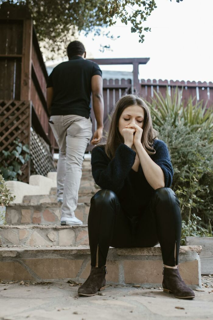 Woman sitting on outdoor steps looking upset while a man walks away, illustrating gender inequality themes.