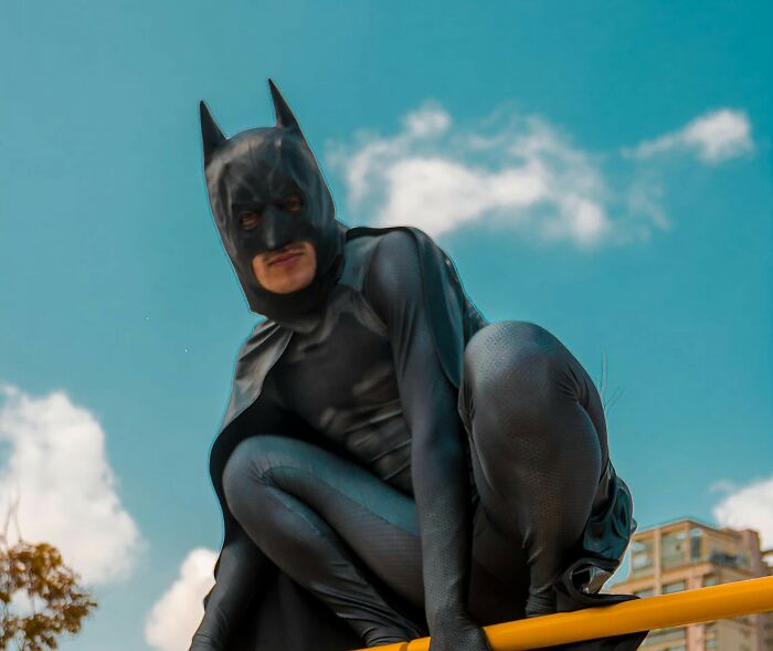 Person dressed as Batman crouching on a yellow bar outdoors against a blue sky showing a school incident theme.