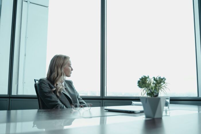 Woman in a gray blazer sitting thoughtfully by office window, reflecting on subtle ways misogyny appears daily.
