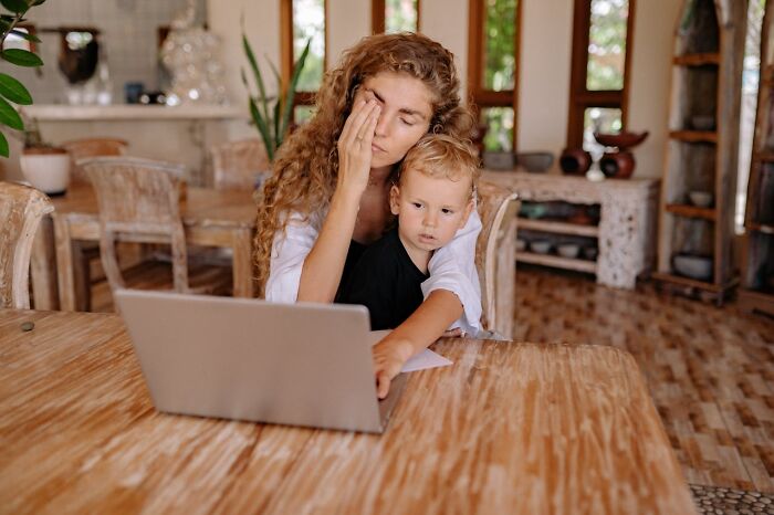 Stressed woman working on laptop while holding child, highlighting challenges women face in work-life balance and freedoms.