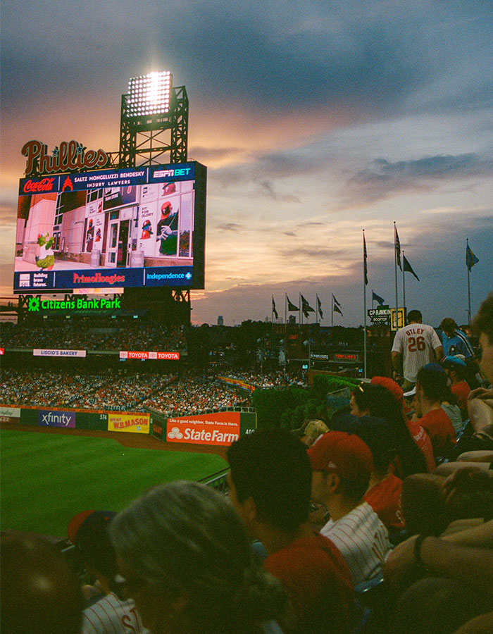Crowd watching a Phillies baseball game at Citizens Bank Park during sunset with scoreboard displaying Phillies mascot. Crowd watching a Phillies baseball game at Citizens Bank Park during sunset with scoreboard displaying Phillies mascot.