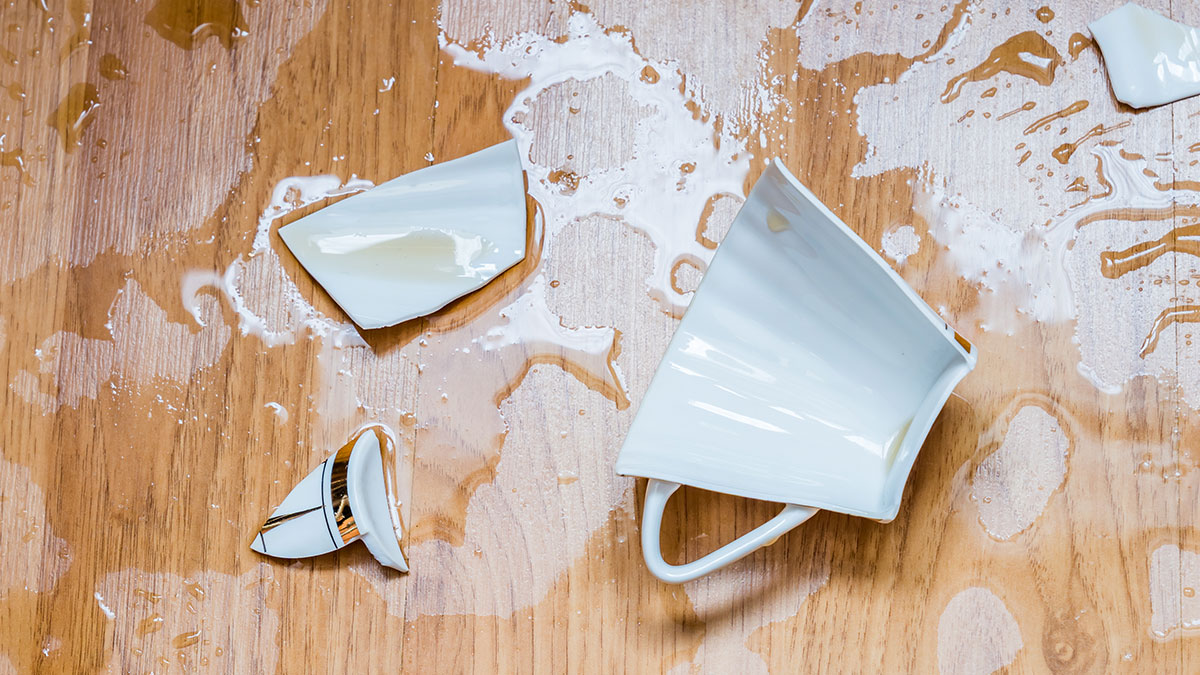 Broken white ceramic cup and spilled liquid on wooden floor illustrating habits people who used to be poor can't get rid of.