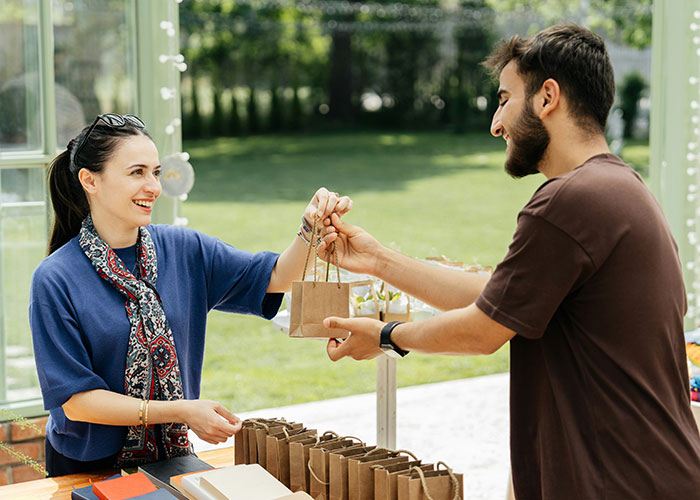 Woman handing a product bag to a smiling man outdoors, illustrating job descriptions where person manipulates another to have a product.