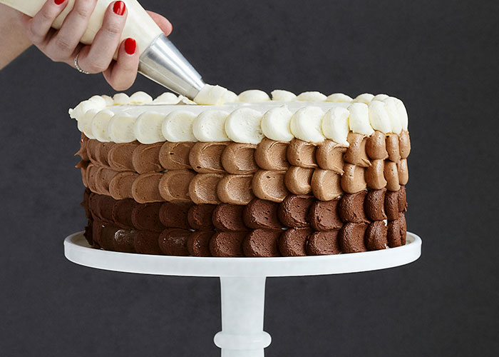 Hand piping white frosting on a multi-layered chocolate cake, showcasing skilled product manipulation techniques.
