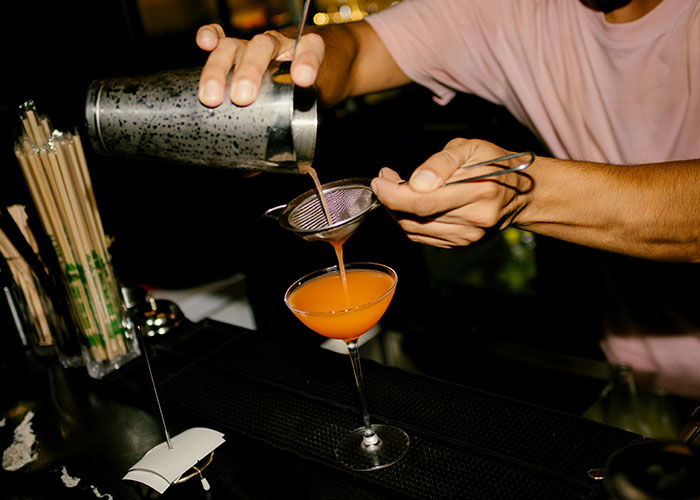Bartender manipulating a person pouring a cocktail through a strainer into a glass in a dimly lit bar.