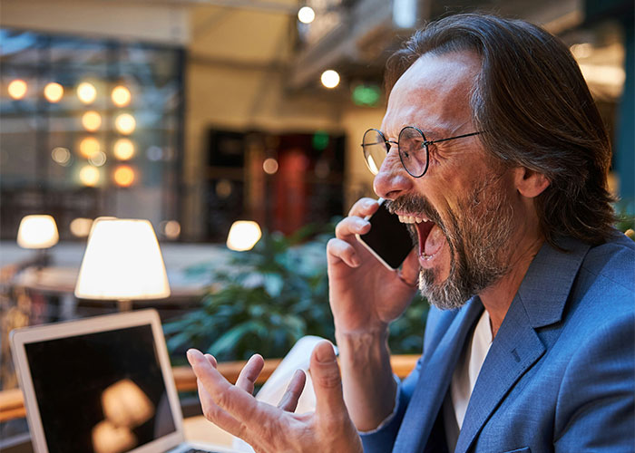 Man in glasses and blue blazer intensely talking on phone at laptop, showing frustration in a modern office setting.