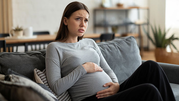 Pregnant woman looking worried on couch risking anaphylaxis as sister-in-law ignores no-peanut allergy rule at home.