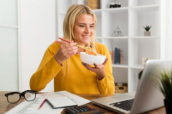Young woman in yellow sweater eating noodles while working on a laptop, illustrating a serial food thief with peanut allergy story. Young woman in yellow sweater eating noodles while working on a laptop, illustrating a serial food thief with peanut allergy story.