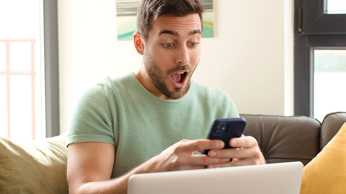 Man in green shirt surprised by inspiring facts on smartphone while sitting on couch with laptop nearby