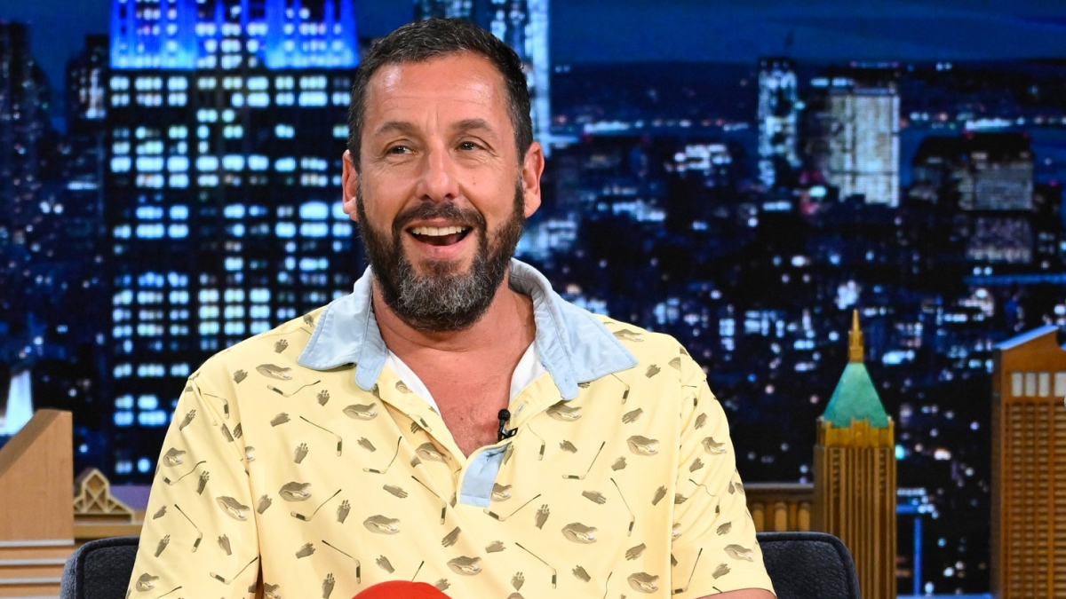 Adam Sandler smiling in a patterned yellow shirt with a light blue collar during a nighttime talk show.