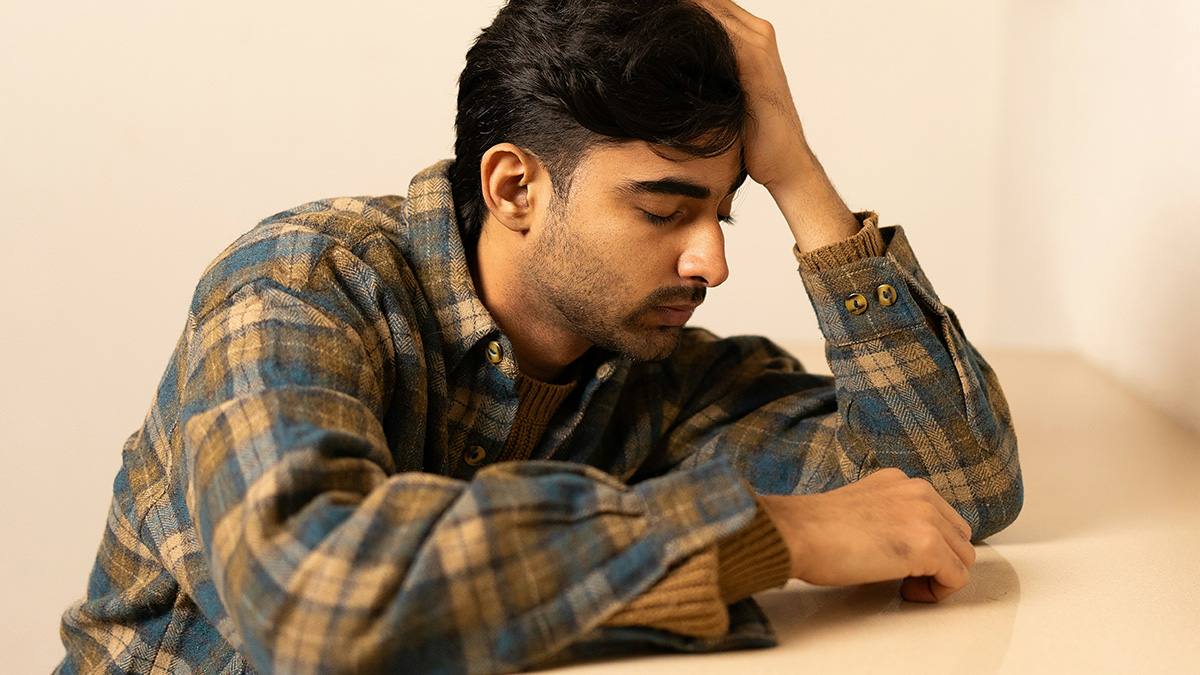 Deaf man sitting quietly with eyes closed and head resting on hand, wearing a plaid jacket indoors.