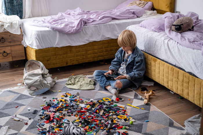 Young boy sitting on a messy bedroom floor with toys scattered, illustrating the teen creates hygiene checklist for bro before school. Young boy sitting on a messy bedroom floor with toys scattered, illustrating the teen creates hygiene checklist for bro before school.