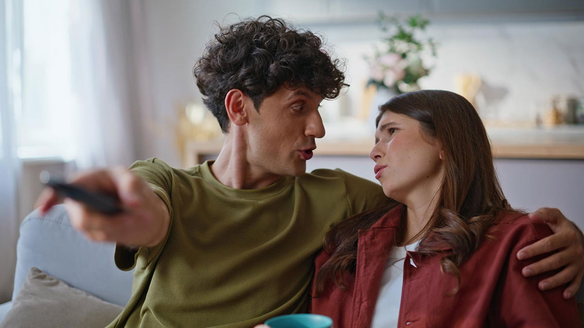 Autistic guy arguing with bestie on couch, holding remote, showing frustration over favorite show disagreement.
