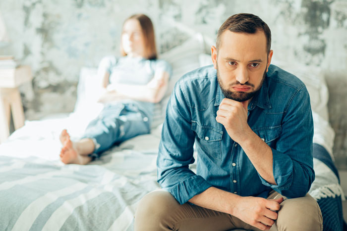 Man looking regretful sitting on bed with partner blurred in background, reflecting on marriage ruined by online affair. Man looking regretful sitting on bed with partner blurred in background, reflecting on marriage ruined by online affair.