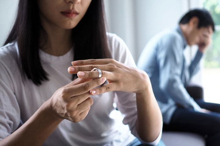 Woman removing wedding ring while man sits in background looking distressed, symbolizing marriage ruined by online affair.