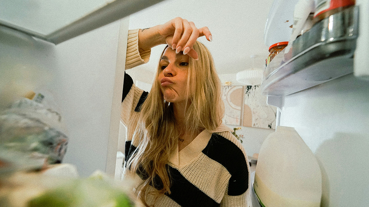 Woman looks disappointed opening a crowded fridge, illustrating lack of space to keep lunch in the fridge at work