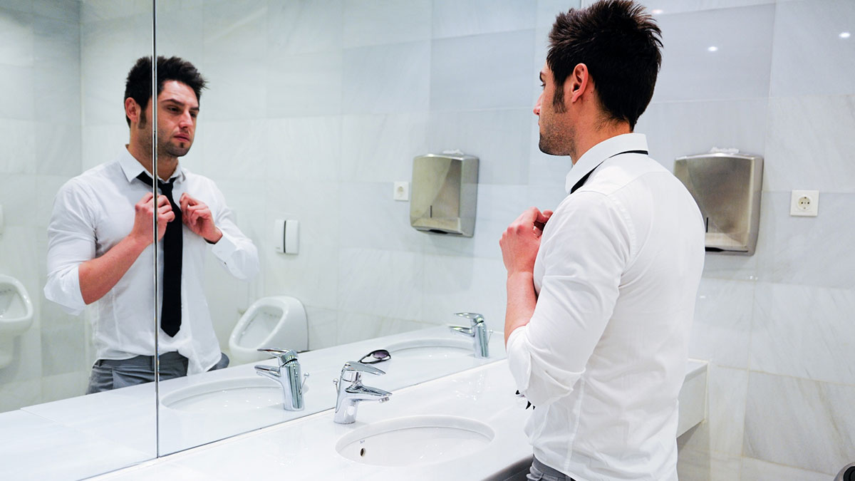 Man in men's restroom adjusting tie, reflecting discomfort, highlighting issues with men's restroom safety and cleanliness concerns.