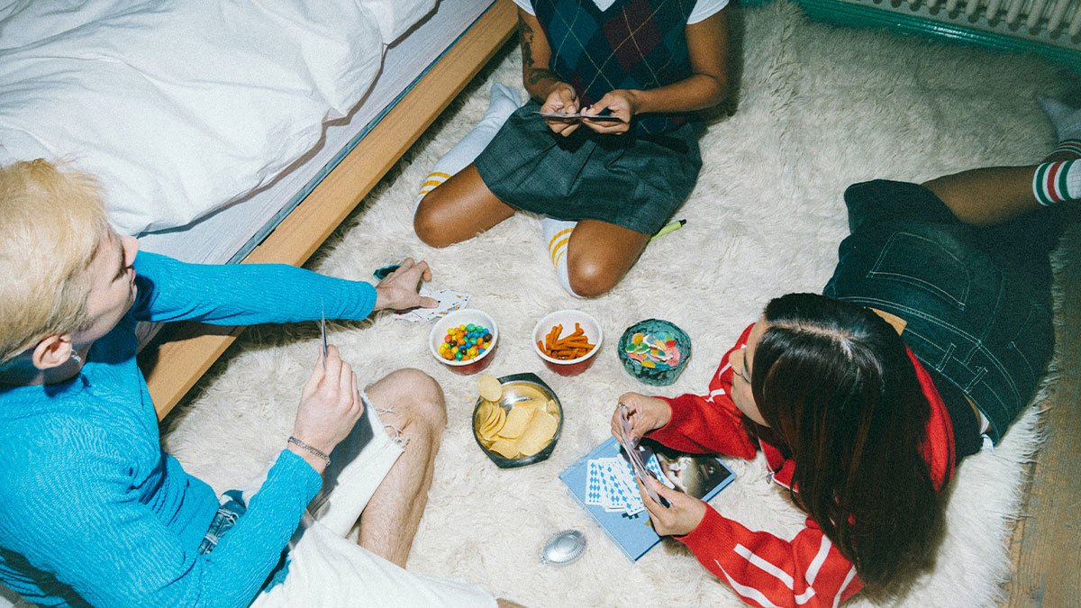 Three friends playing cards on a rug with snacks nearby, capturing a social setting with boxers and casual wear.