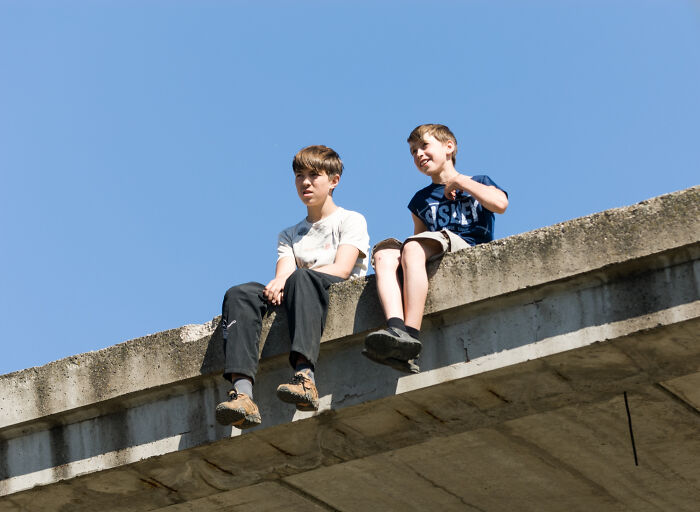 Two boys sitting on the edge of a concrete structure under clear blue sky, reflecting babysitters horror experiences.