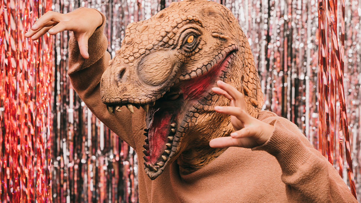 Person wearing a dinosaur mask and brown sweater, posing playfully against a shiny red and silver backdrop for a prank.