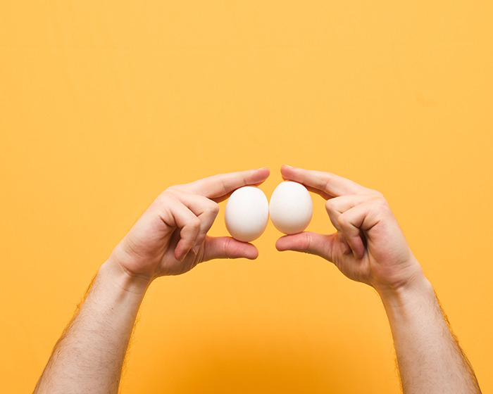 Two hands holding white eggs against a yellow background representing the concept of Scrotox cosmetic trend among men. Two hands holding white eggs against a yellow background representing the concept of Scrotox cosmetic trend among men.
