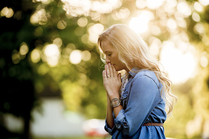 Young woman in denim jacket praying outdoors, evoking themes of real-life hexes, curses, and witchcraft beliefs.
