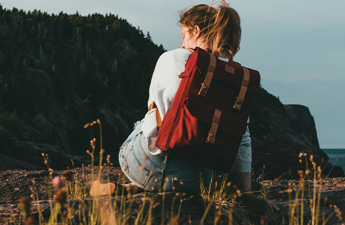 Teen girl with a red backpack sitting on a rock overlooking nature, reflecting on school gossip turned to facts.