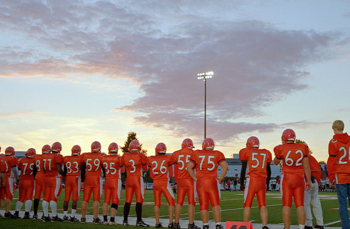 High school football players in orange uniforms standing on the sidelines at sunset, illustrating school gossip and facts.
