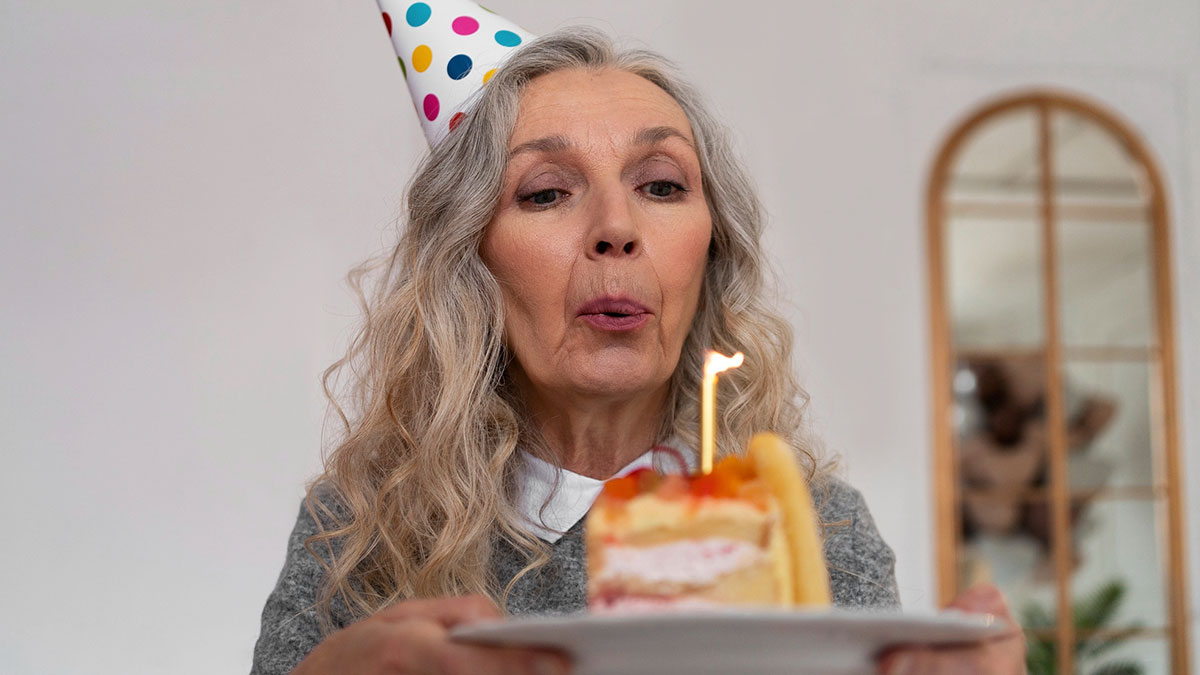 Older woman wearing a party hat blowing out a candle on birthday cake, symbolizing surprising family secrets uncovered.