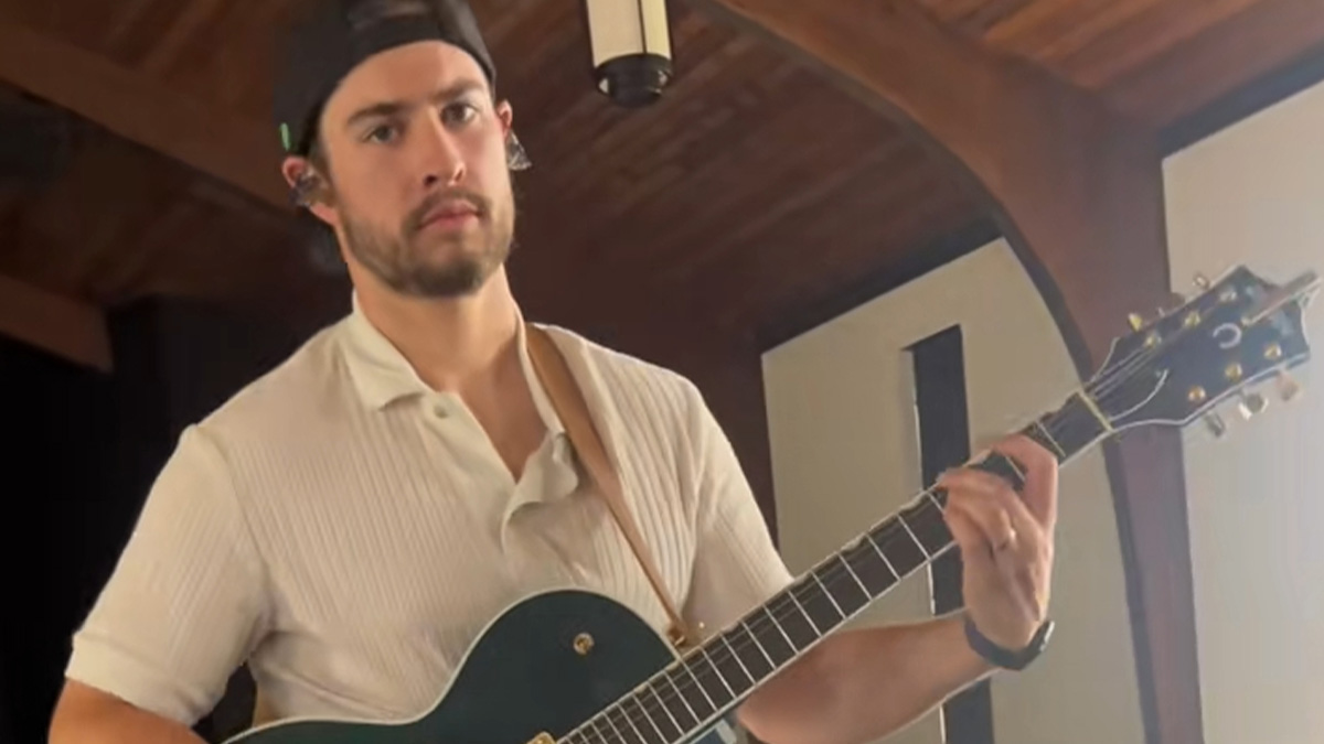 Man playing guitar indoors, wearing a white shirt and cap, showcasing a husband glow-up in a casual setting.