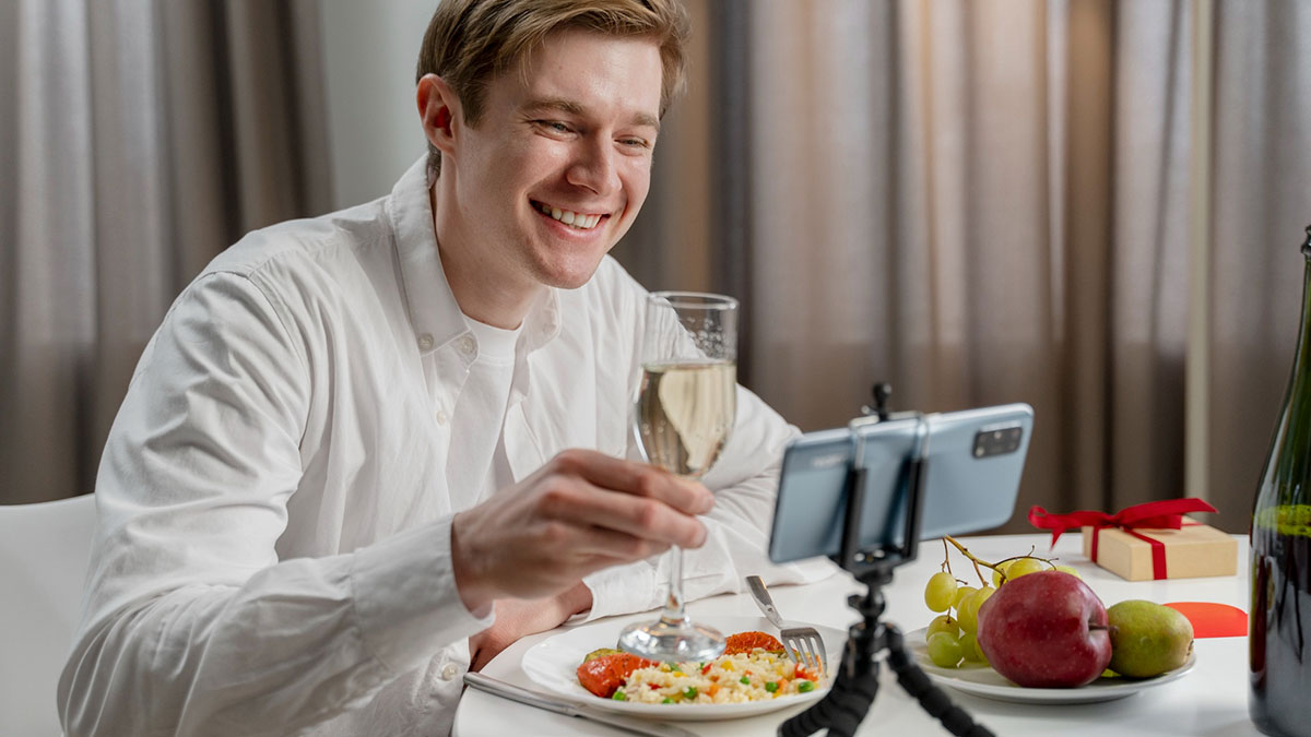 Man smiling during a virtual dinner date, raising a glass while using a smartphone to connect online.