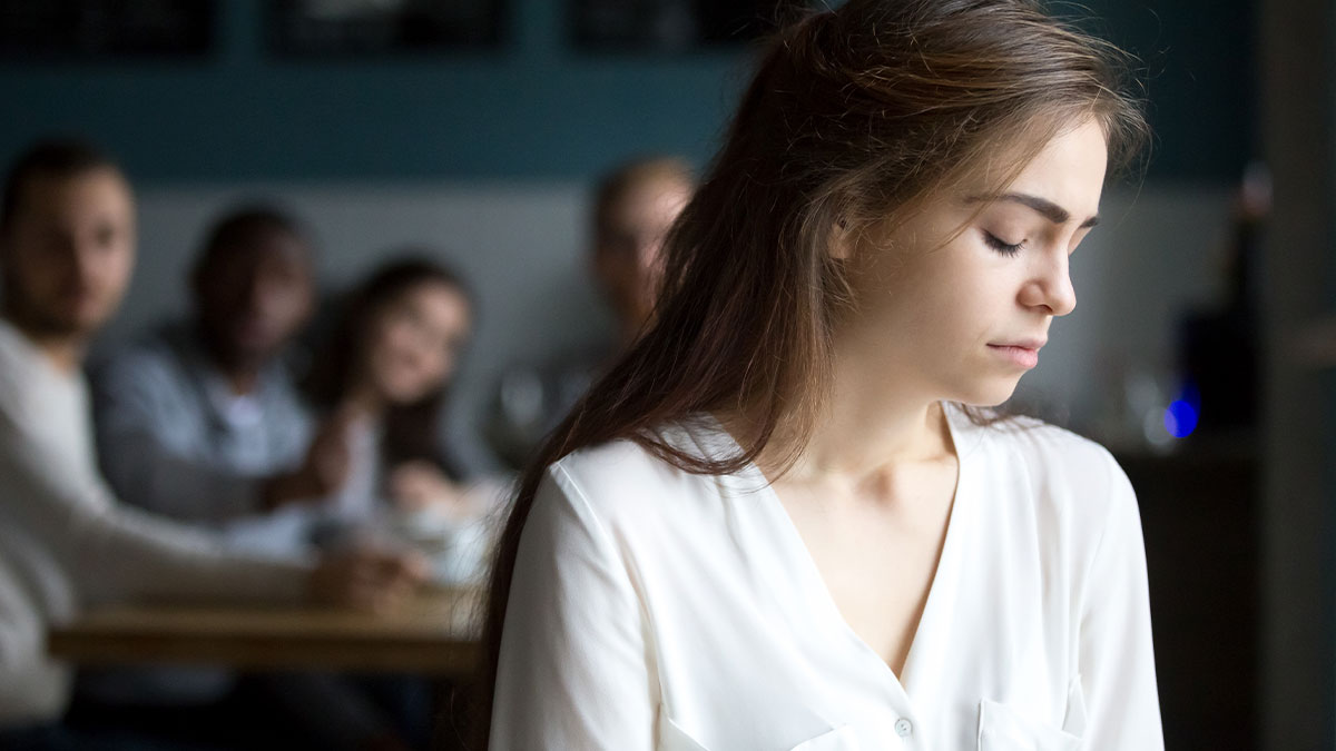 Young woman in white shirt looking down, appearing conflicted as others sit blurred in the background at a table.