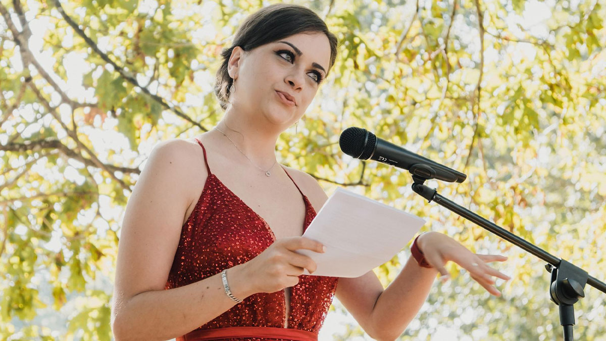 Bride in red dress speaking into microphone outdoors, illustrating family favoritism and twin wedding tension.