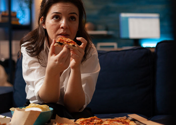 Woman sitting on couch eating pizza, reflecting on body image and emotional impact after learning husband's thoughts. Woman sitting on couch eating pizza, reflecting on body image and emotional impact after learning husband's thoughts.