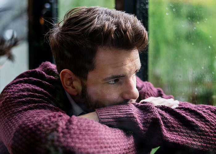 Man in a maroon sweater looking thoughtfully out a window, reflecting on feelings about body and relationship issues. Man in a maroon sweater looking thoughtfully out a window, reflecting on feelings about body and relationship issues.