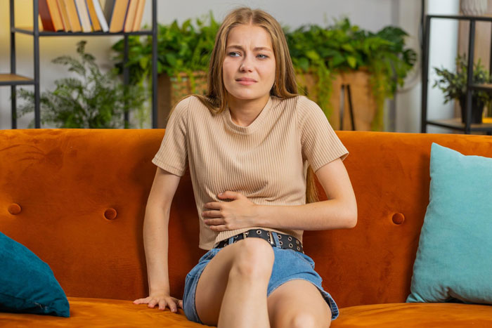 Young woman sitting on a couch looking sick and distressed, possibly affected by parents' food causing illness and anxiety.