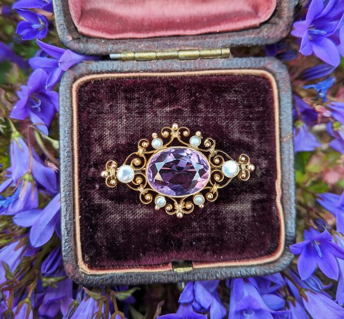 Antique gold brooch with amethyst and pearls in a velvet box surrounded by purple flowers, related to butcher and gravedigger professions.