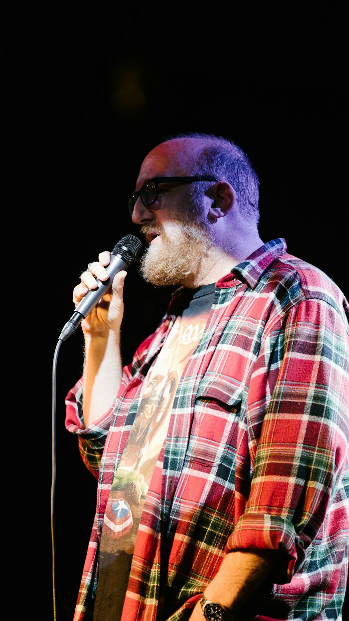 Bearded man in glasses and plaid shirt holding microphone, speaking on stage about things women are not allowed to do.